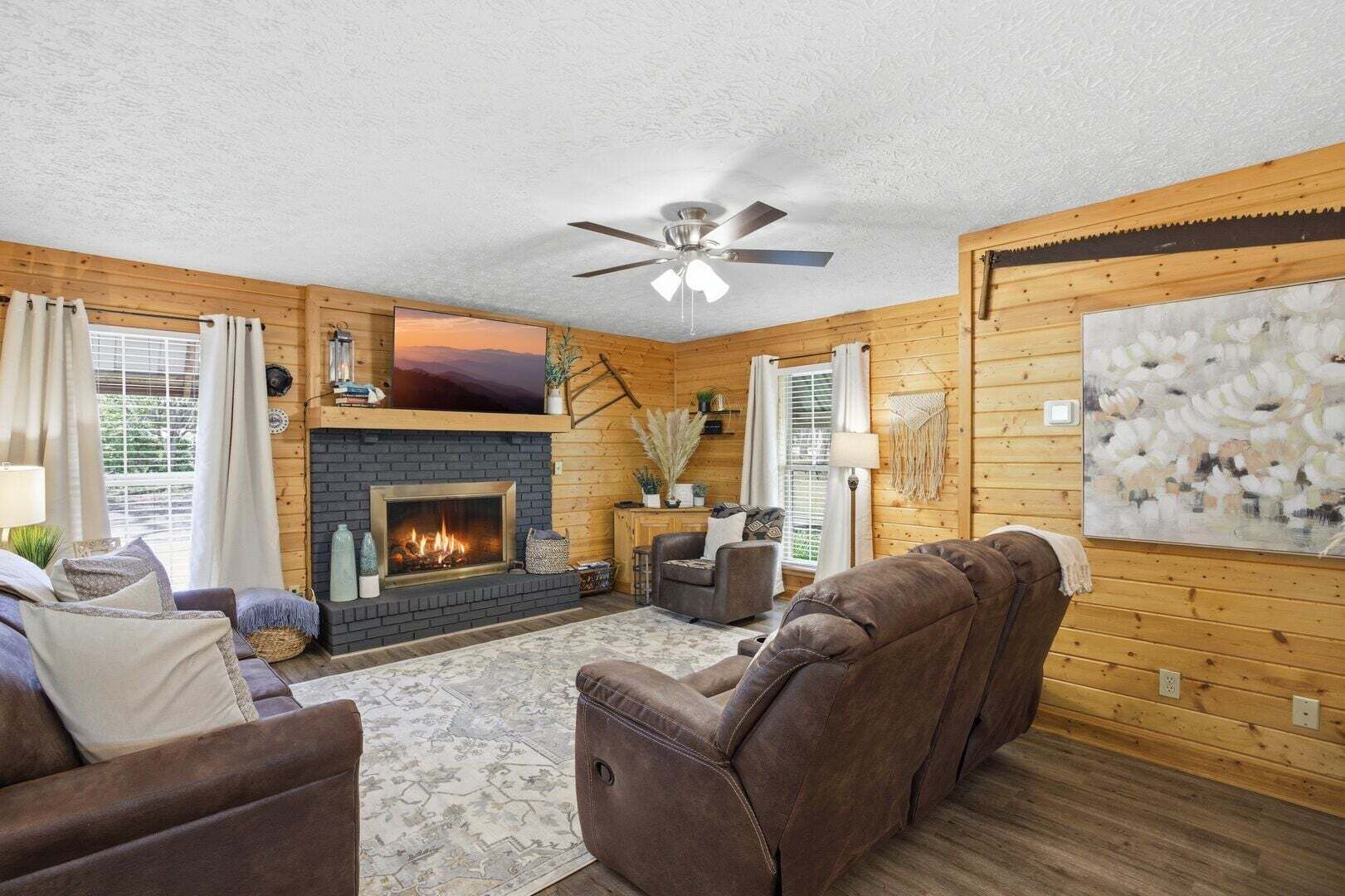 A cozy living room with a fireplace, wooden walls, and a ceiling fan.