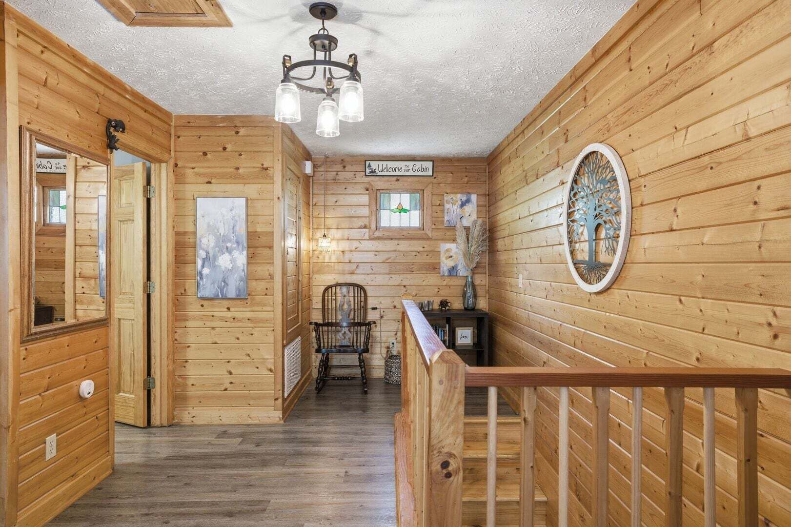 A wooden cabin interior with a staircase, a welcome sign, a chair, and a decorative mirror.