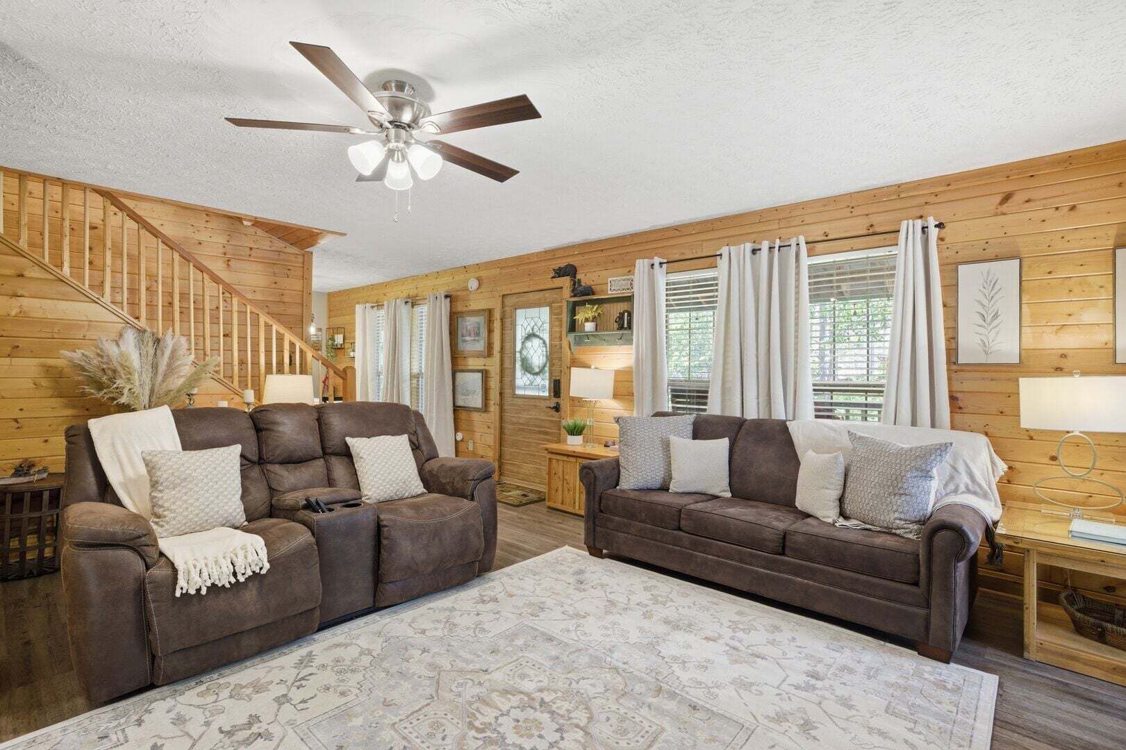 A log cabin interior with two leather sofas, a patterned rug, a ceiling fan, wooden walls, and a staircase.