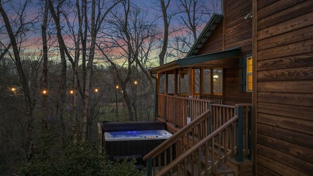 Una cabaña de madera con un jacuzzi en la terraza, rodeada de árboles y un atardecer al fondo.