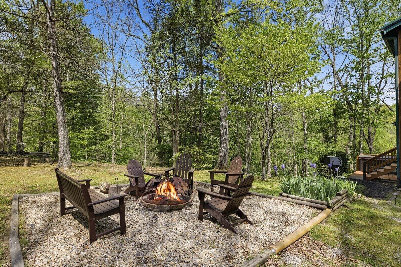 Ein Feuerkorb mit vier Adirondack-Stühlen, umgeben von Kies, in einer bewaldeten Gegend mit Bäumen und einer Holzterrasse.