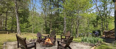 Ein Feuerkorb mit vier Adirondack-Stühlen, umgeben von Kies, in einer bewaldeten Gegend mit Bäumen und einer Holzterrasse.