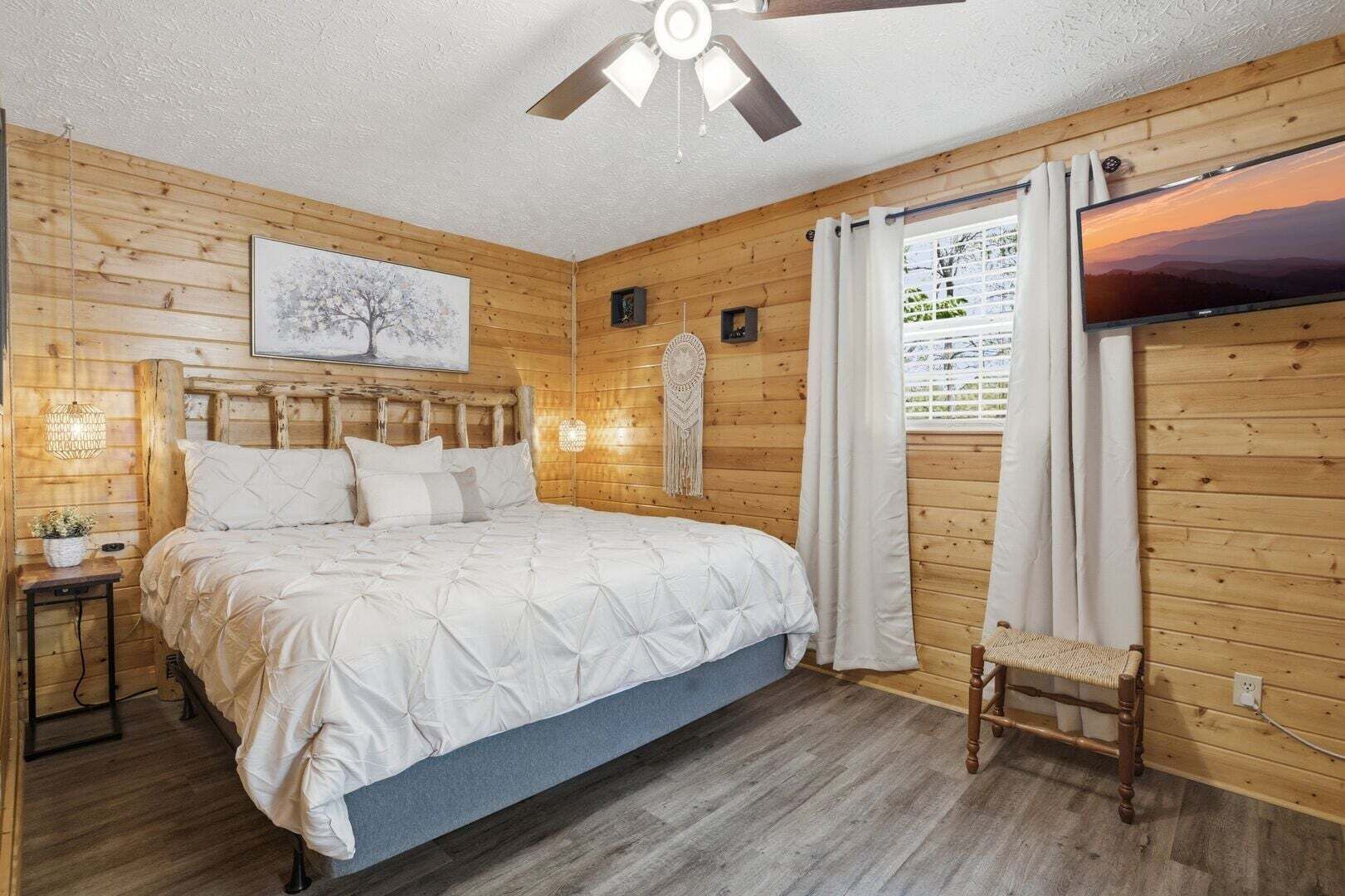 A wooden-paneled bedroom with a bed, a ceiling fan, a wall-mounted TV, and a window with curtains.