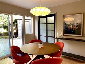 Un coin repas avec une table ronde, des chaises rouges, un luminaire suspendu jaune, une porte vitrée et un tableau abstrait encadré accroché au mur.