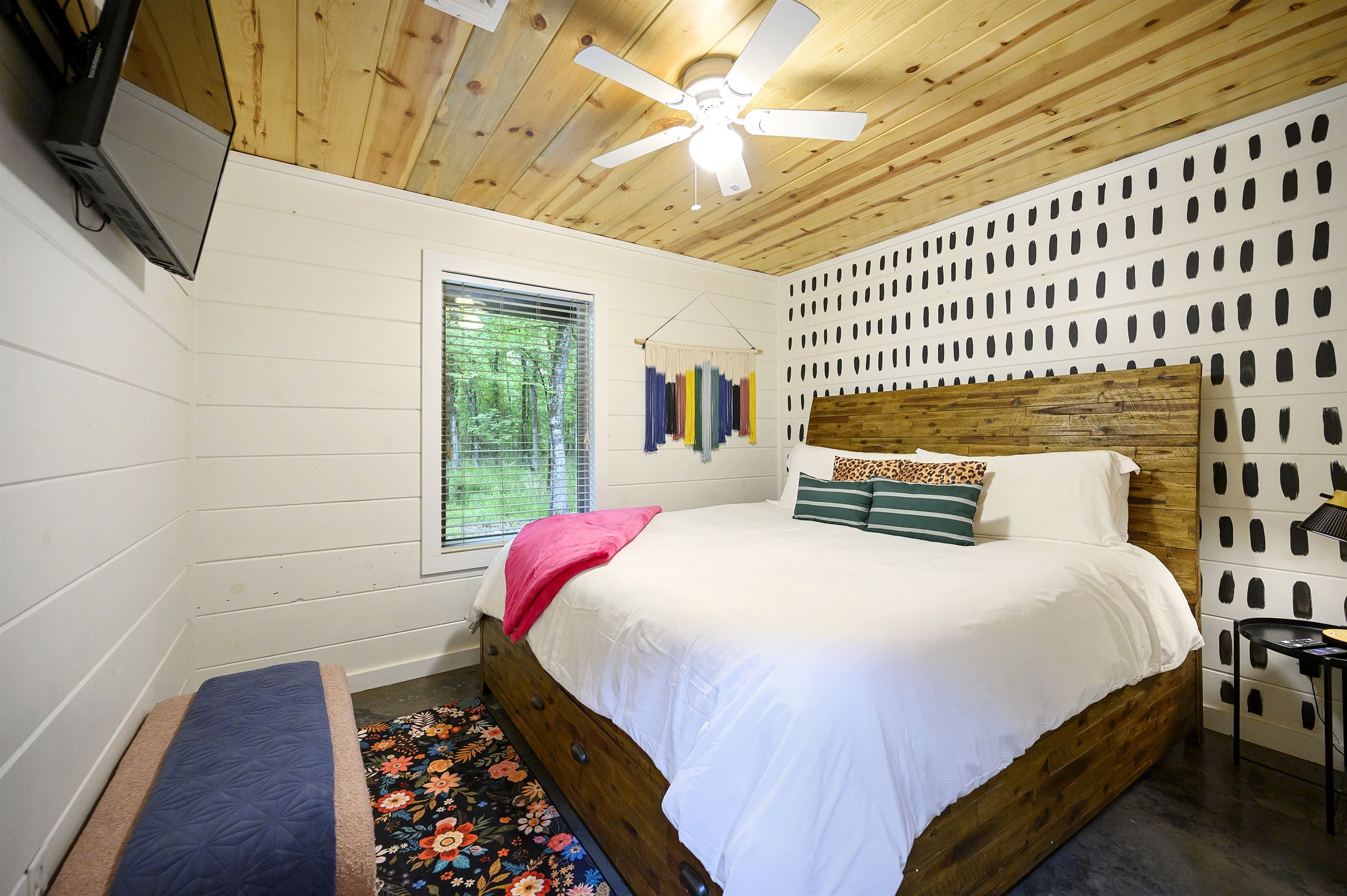 A bedroom with a wooden ceiling, a bed with white bedding, a bench, a window with blinds, and a wall-mounted TV.