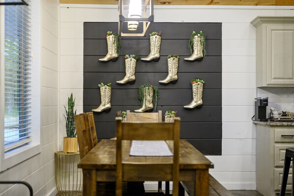 A dining area with a wooden table and chairs, a blackboard wall with decorative boots and plants, a window with blinds, and a kitchen area with cabinets and appliances.