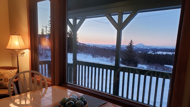 A dining area with a table, chairs, a lamp, and a view of a snowy landscape through a large window.
