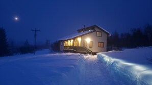 A snow-covered landscape with a house featuring a porch and a staircase leading to the entrance.