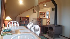 A cozy dining area with a table, chairs, a lamp, a bookshelf, and a wood-burning stove.