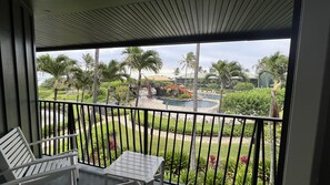 Ein Balkon mit Blick auf eine angelegte Grünfläche, darunter ein Pool, Palmen und ein Gebäude mit Gründach.