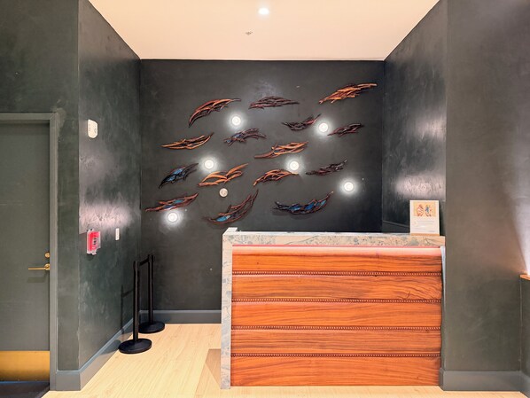 A reception area with a dark wall, a wooden reception desk, and a series of wall-mounted decorative elements. - Kauai Beach Resort, Top Floor, Pool View (Kauai)