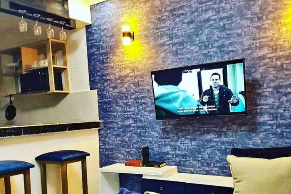 A modern living room with a flat-screen TV mounted on the wall, a blue sofa, and two wooden stools.