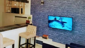 A modern living room with a flat-screen TV mounted on a textured wall, a sofa, two bar stools, and a wooden coffee table. - Sandalwood Suites Nakuru (Nakuru)