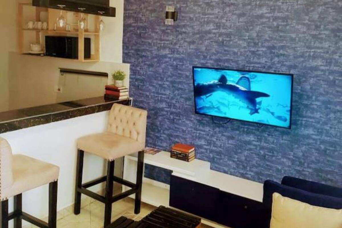 A modern living room with a flat-screen TV mounted on a textured wall, a sofa, two bar stools, and a wooden coffee table.