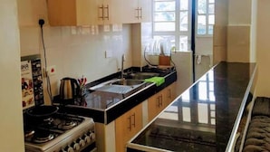 A kitchen with a stove, oven, and black countertop. There is a window with blinds and a door in the background. - Sandalwood Suites Nakuru (Nakuru)