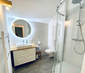 Un baño moderno con cabina de ducha de vidrio, un espejo redondo, un lavabo blanco y un inodoro.