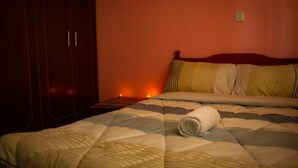 A bedroom with a bed, a wooden wardrobe, and a bedside table with a lamp. - Fountain Apartments (Naivasha)