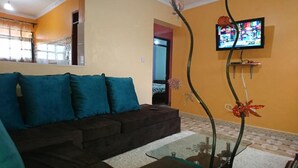 A living room with a dark brown sofa, teal cushions, a glass coffee table, and a television mounted on the wall. - Fountain Apartments (Naivasha)