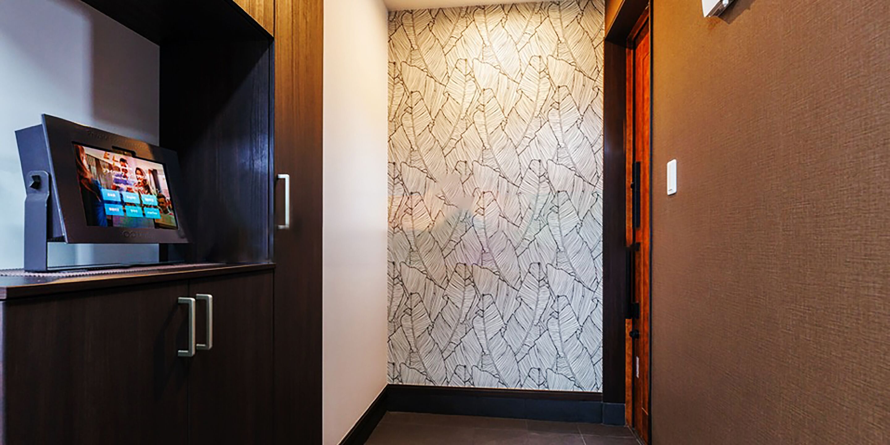 Un pasillo de hotel con una pantalla digital en un mueble, papel tapiz estampado y una puerta de madera.