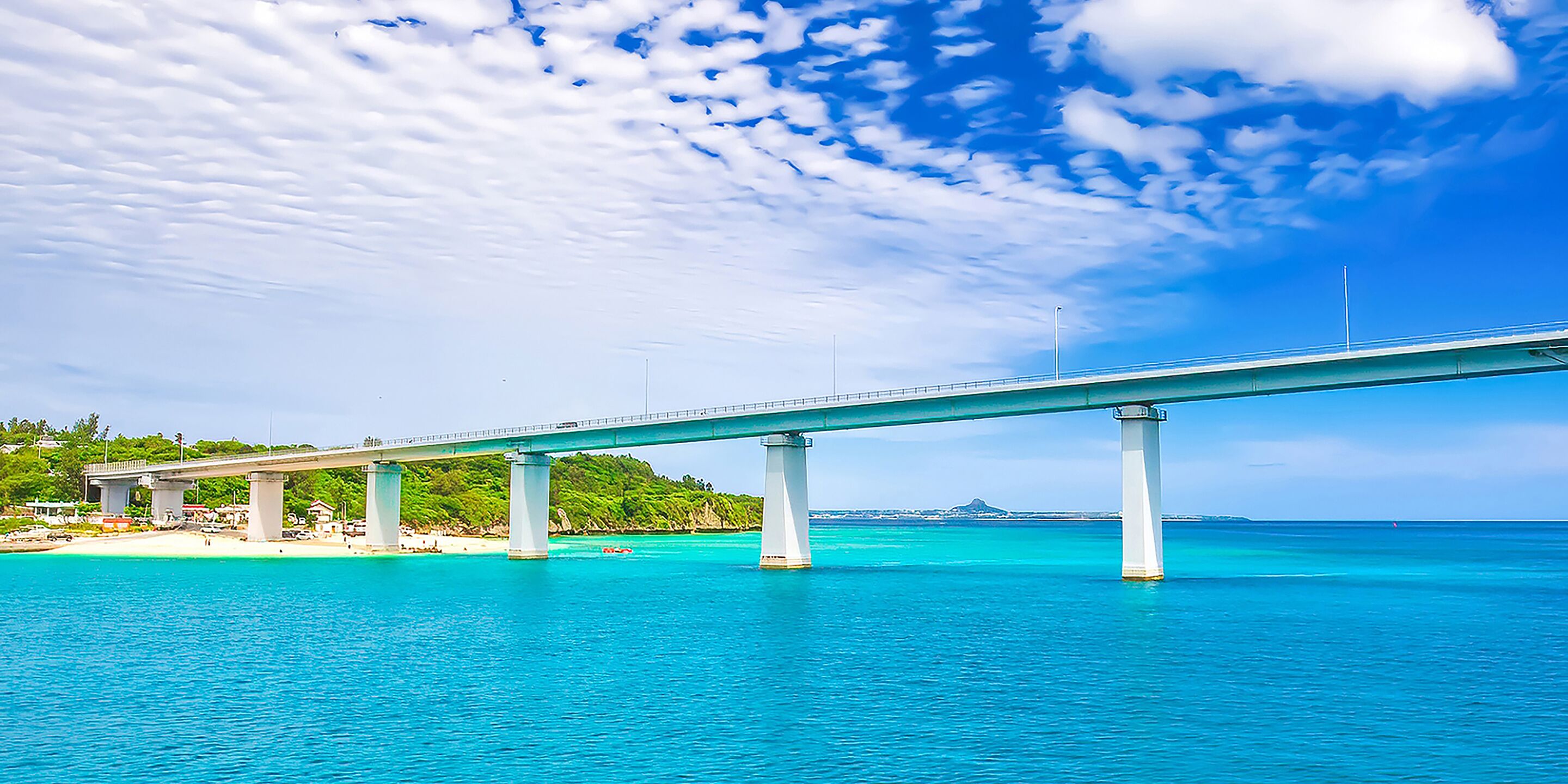 A bridge over turquoise water with a beach and greenery in the background.