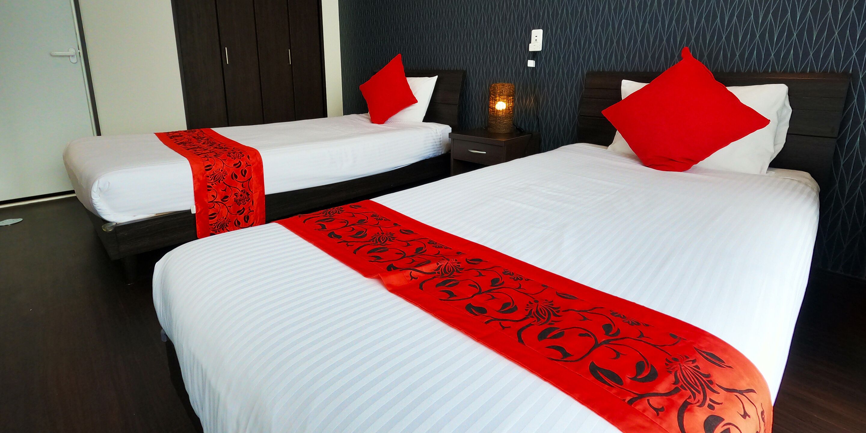 Two double beds with red and black bedspreads, a nightstand with a lamp, and a dark wooden wardrobe.