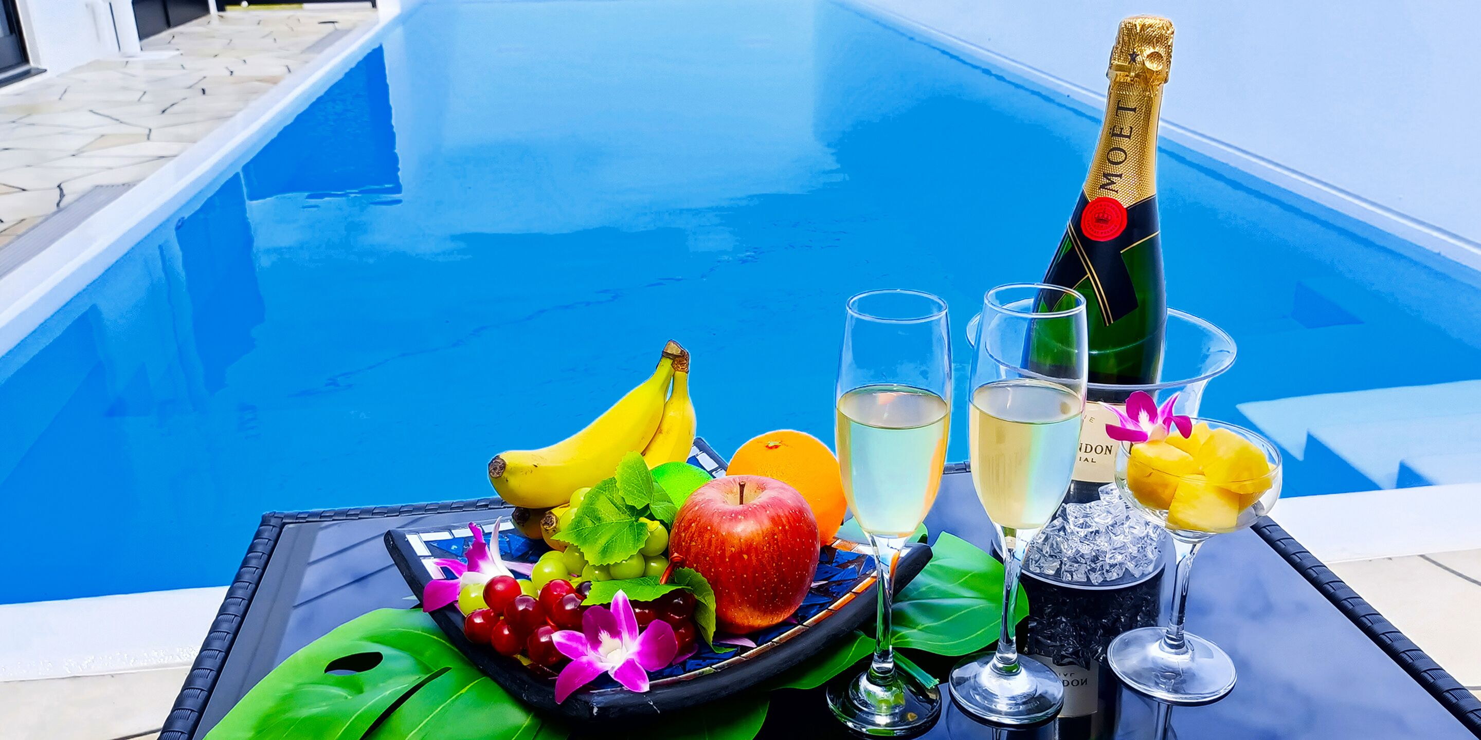 A poolside table with a bottle of champagne, two glasses, a fruit platter, and a bowl of ice.