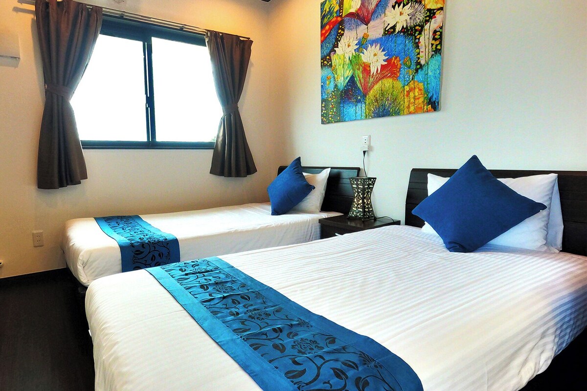 A hotel room with two beds, a window with curtains, a painting on the wall, and a nightstand with a lamp.