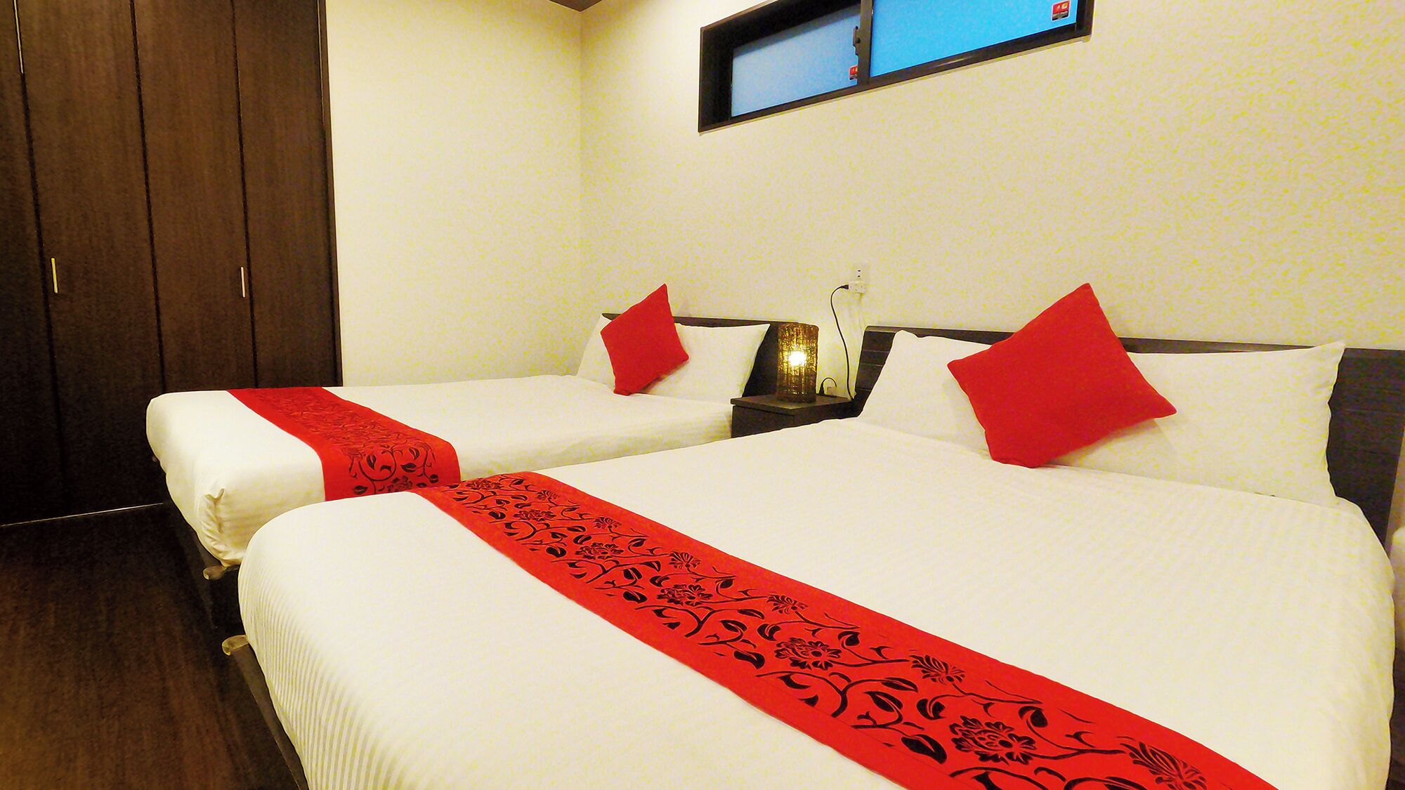 A hotel room with two beds, each with a red decorative runner, white bedding, and red pillows.