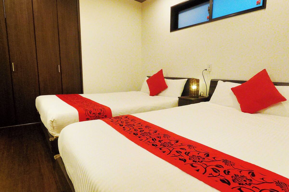 A hotel room with two beds, each with a red decorative runner, white bedding, and red pillows.