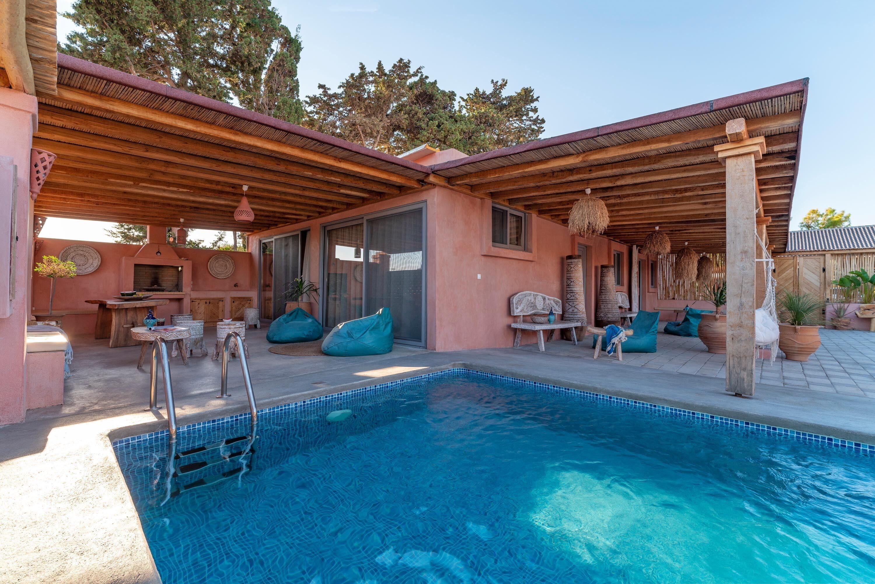 Zona de piscina con una pérgola de madera, asientos y una piscina azul cristalina.