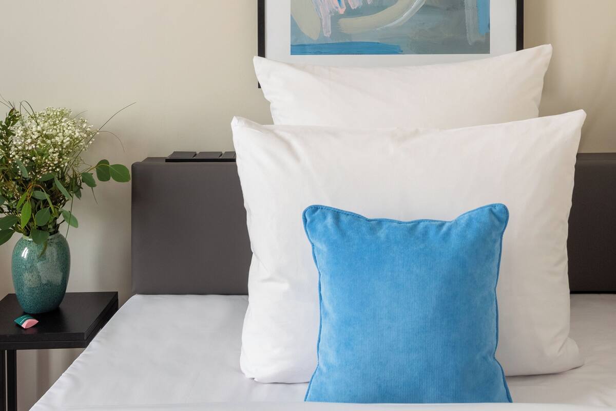 A neatly made bed with white and blue pillows, a framed abstract painting above it, and a small vase with white flowers on a nightstand beside the bed.