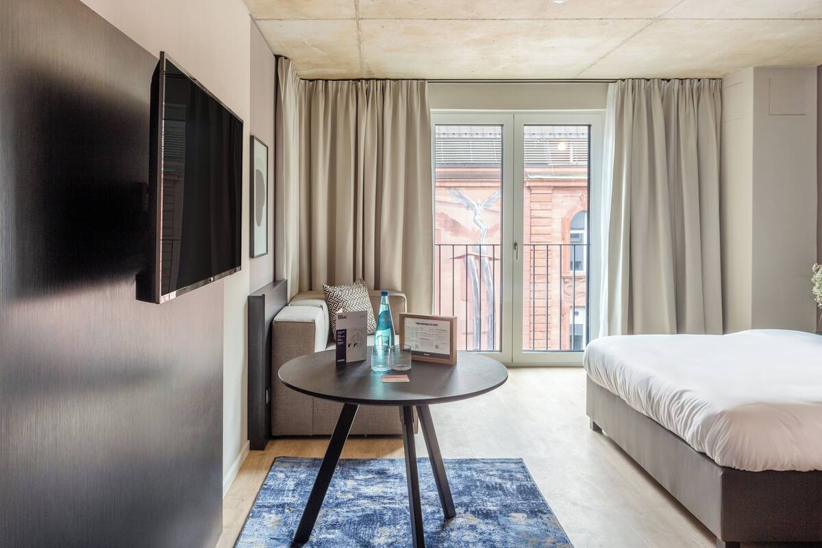 A hotel room with a bed, a round table with a chair, a TV mounted on the wall, and a balcony with curtains.