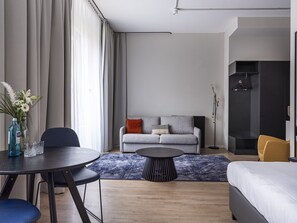 Ett modernt hotellrum med en grå soffa, ett runt soffbord, en säng och en matplats med ett bord och stolar.