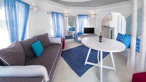 A modern living room with a sofa, a round table, and a TV. There is a kitchen area visible through an open door. - Kouros Home and Suites (Rhodes)