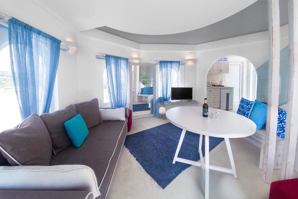 A modern living room with a sofa, a round table, and a TV. There is a kitchen area visible through an open door.