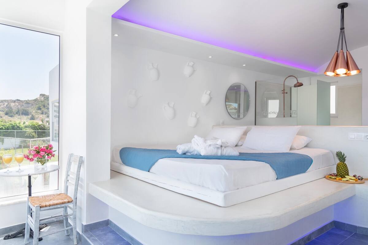 A modern bedroom with a curved bed, a small table with drinks, a chair, and a balcony view.