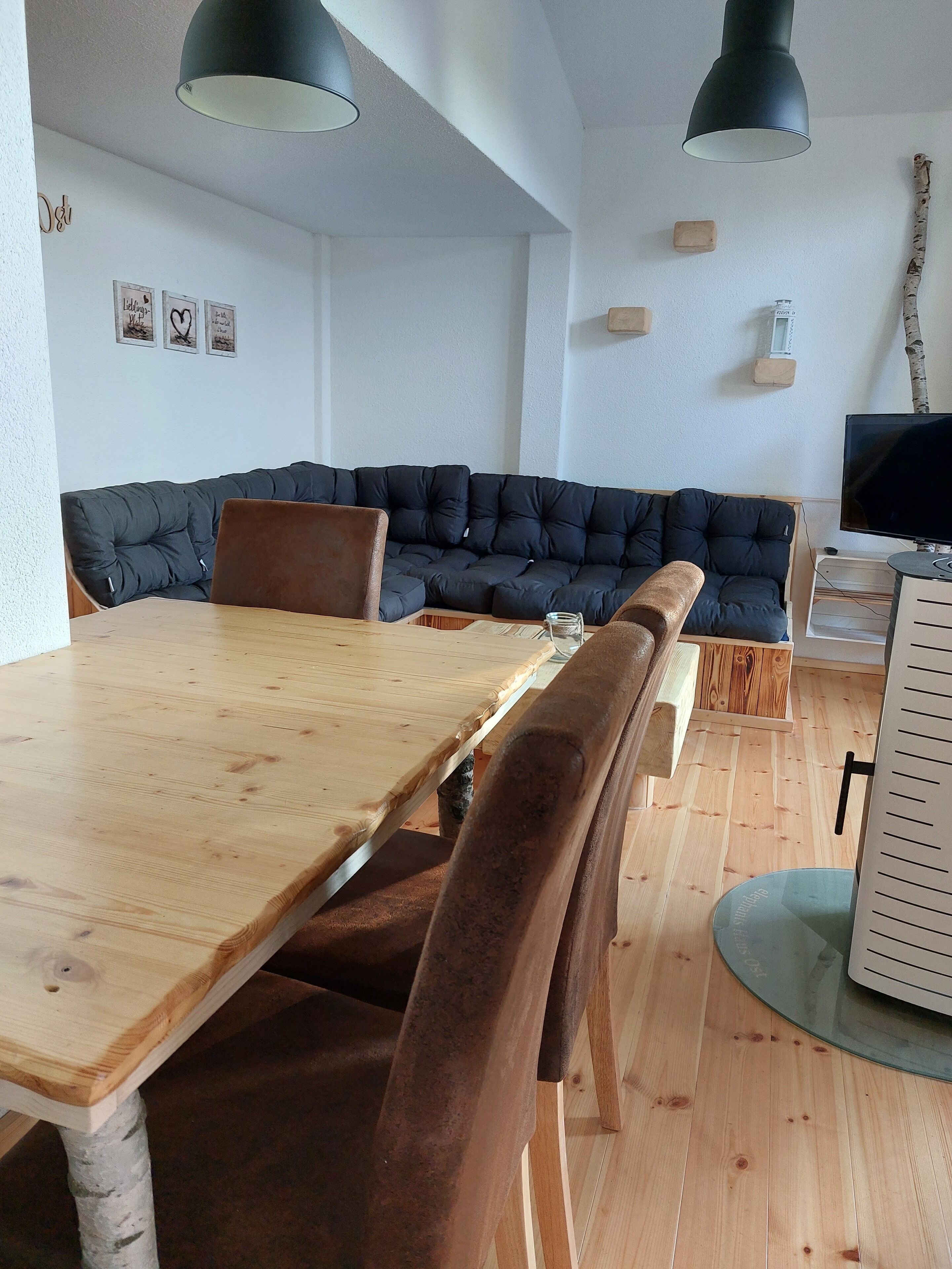 A modern living room with a wooden dining table, brown cushioned chairs, a dark blue sofa, and two black pendant lights.