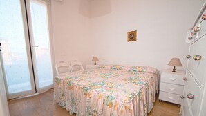 Un dormitorio con colcha estampada, muebles blancos, una mesita de noche con lámpara y un mueble de pared con pomos decorativos.