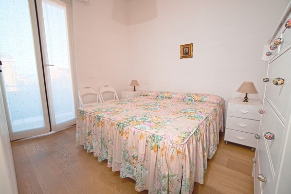 Ein Schlafzimmer mit floralem Bettbezug, weißen Möbeln, einem kleinen Nachttisch mit Lampe und einem an der Wand befestigten Schrank mit dekorativen Knöpfen.