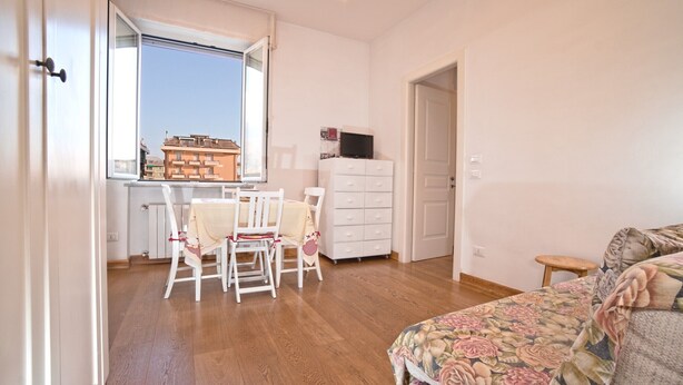Una habitación con piso de madera, una cama con ropa de cama floral, un tocador blanco, un televisor encima y una ventana con vista a edificios.