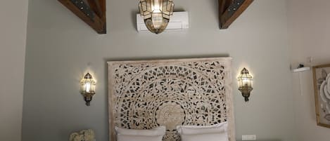 A bedroom with a wooden ceiling, a large bed with a decorative headboard, two wall-mounted lamps, and a bedside table with a vase of flowers.