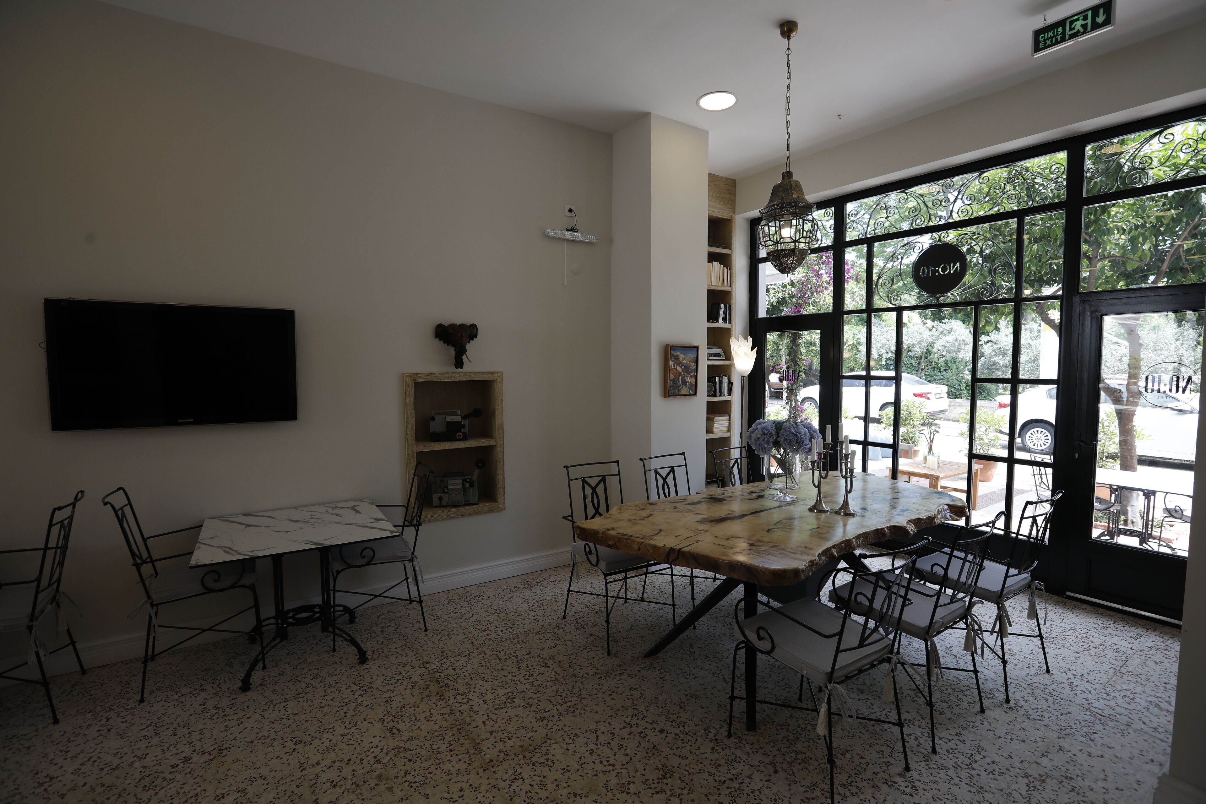 A dining area with a large wooden table and chairs, a television mounted on the wall, and a view of the outside through large windows.
