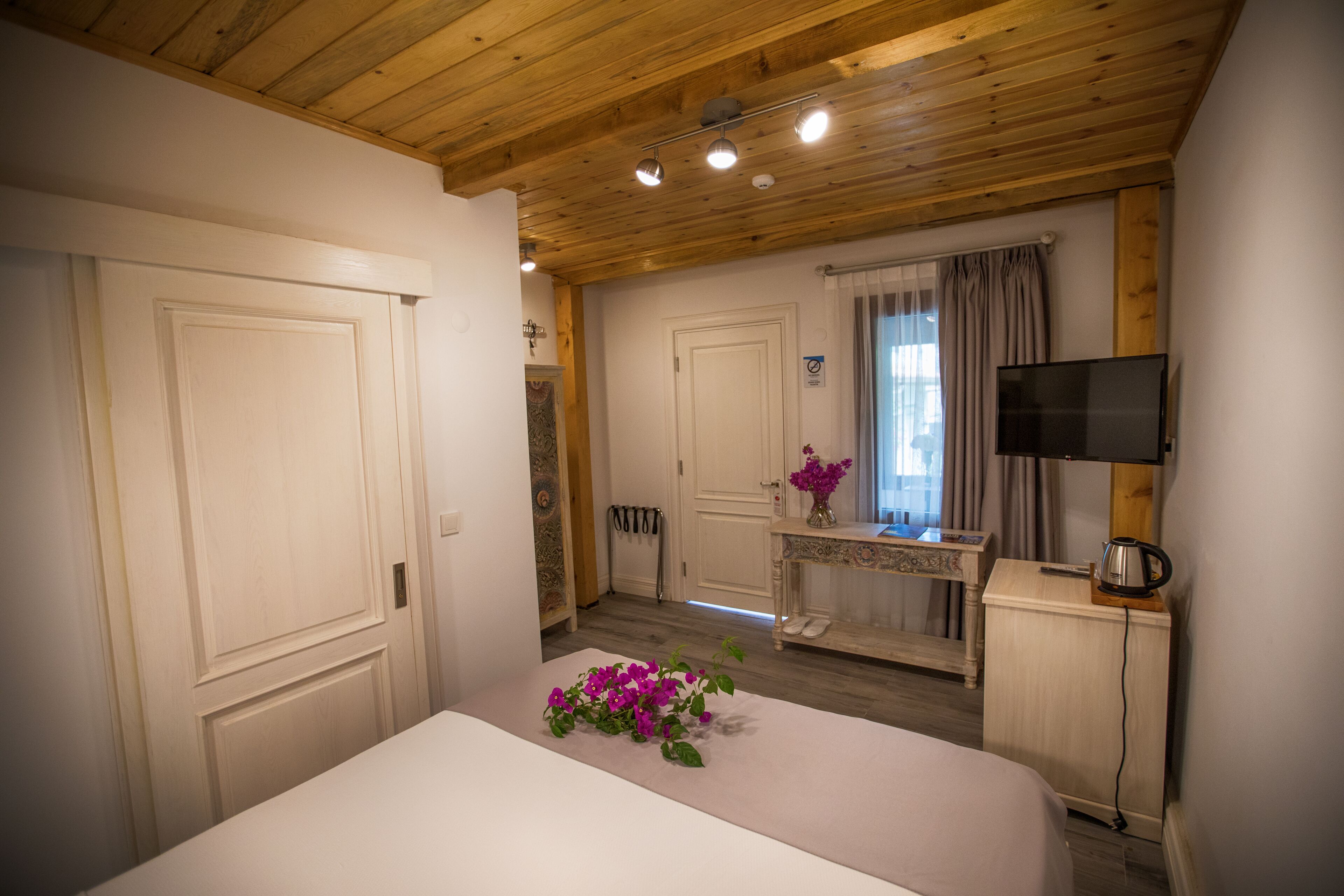 Habitación de hotel con techo de madera, una cama, un escritorio con televisor, un armario pequeño y una ventana con cortinas.