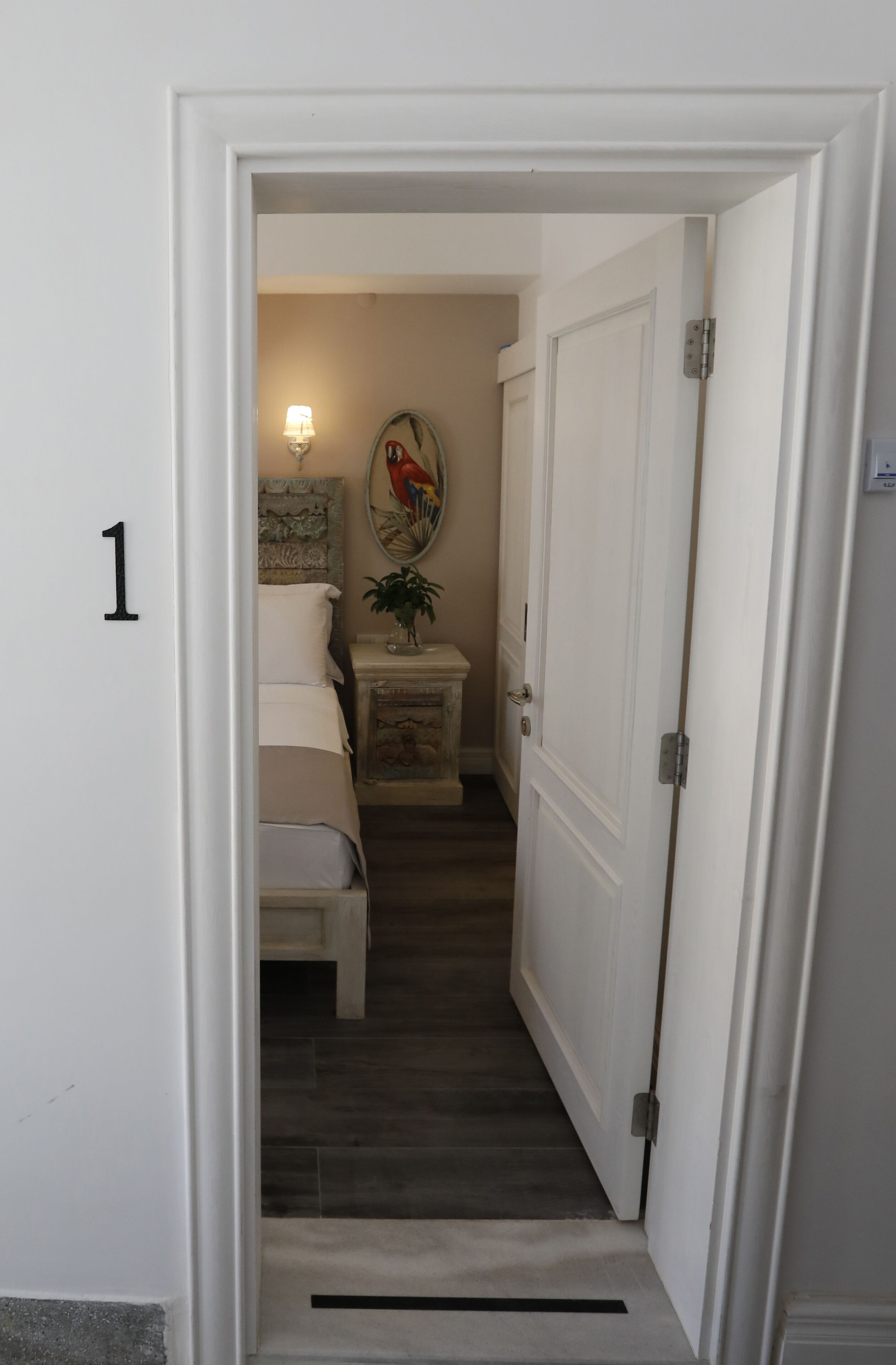 Un pasillo estrecho con una cama, una mesita de noche, una lámpara de pared y una puerta.