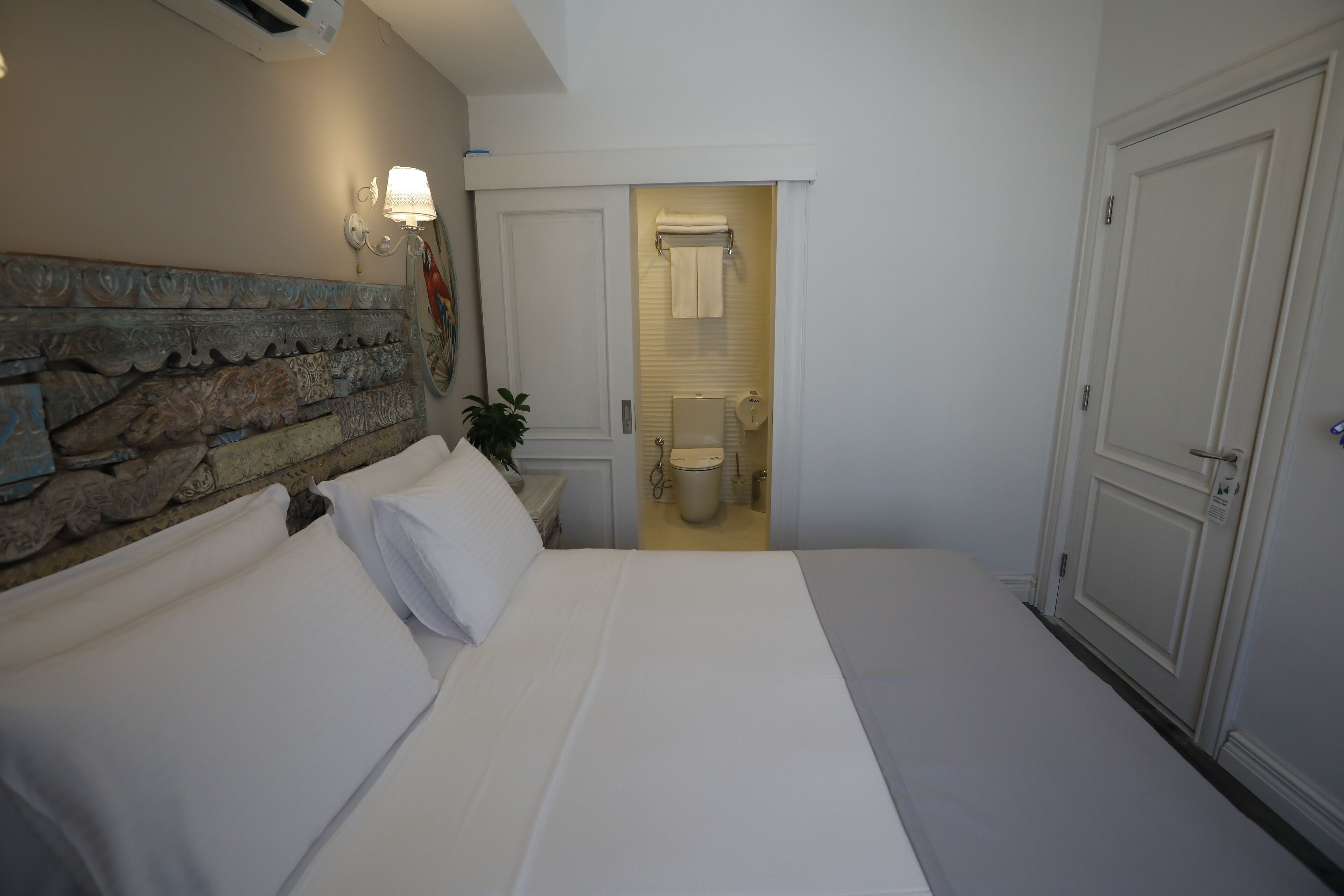Quarto de hotel com uma cama grande, cabeceira decorativa, mesa de cabeceira com abajur e vista para o banheiro.