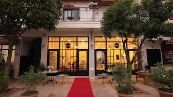A hotel entrance with a red carpet, potted plants, and a balcony with flowers.