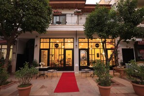 Entrada de hotel con alfombra roja, plantas en macetas y un balcón con flores.