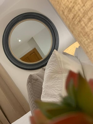 A room with a round mirror on the wall, a bed with a white pillow, and a plant in the foreground. - Cheerful 2-bed Cottage With 2 Free Parking, Leeds (Leeds)