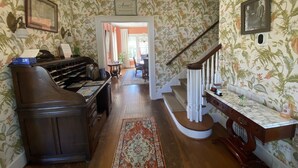 Une pièce avec un bureau en bois, un piano, un escalier et un papier peint à motifs floraux.
