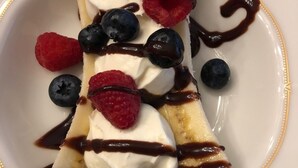 A dessert plate with banana slices, whipped cream, blueberries, raspberries, and chocolate sauce.
